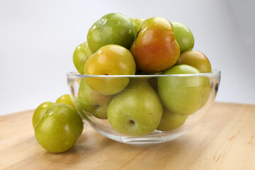 Small mini green fresh juicy plum fruit in transparent glass bowl wooden table white background