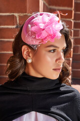 Cropped close-up shot of a young woman in a pink felt pillbox hat decorated with a flower, beads and feathers. A girl with curly hair in the hat is on a brick wall. Front view.