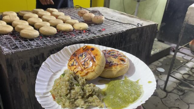 In the backdrop, a vendor is seen baking the Litti in a hot iron grill while serving it with Baigan Bharta and green in a paper plate