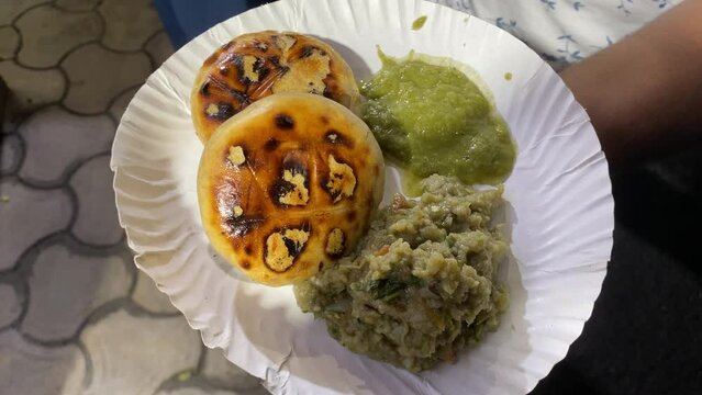 Video depicts a man holding a plate of savory litti chokha smeared with ghee and served with roasted mashed eggplant or baingan bharta and a green spicy chutney.