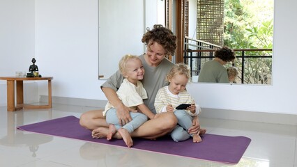 A young mother of twin girls sits with them on the floor together and hugs them. They recently finished doing yoga. Now everyone is happy together and watching something on a mobile phone.