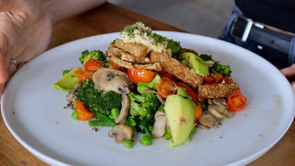 On a white plate, top view, there is a portion of fresh salad of vegetables, mushrooms, meat. A woman moves her fork in her plate, choosing what to eat first. Healthy and delicious food.