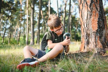 schoolboy is exploring nature with magnifying glass. Summer vacation for inquisitive kids in forest. Hiking. Boy-scout.