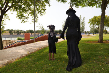 Obraz premium Happy halloween. Child dressed up for halloween party and his mother dressed as a witch walking through the park seen from behind. Trick or treat. 31st October