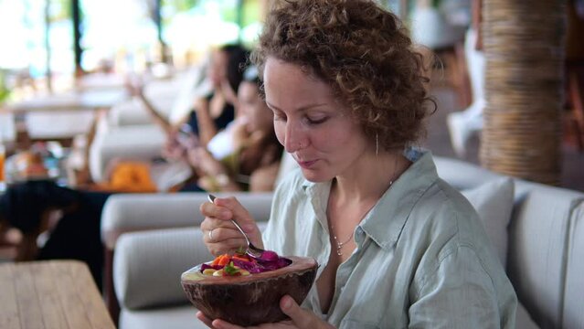 A beautiful woman eats an appetizing dish with fruits and healthy porridge in coconut shell plates in a vegan restaurant. Healthy fruit breakfast, food to fill you with energy for the whole day.