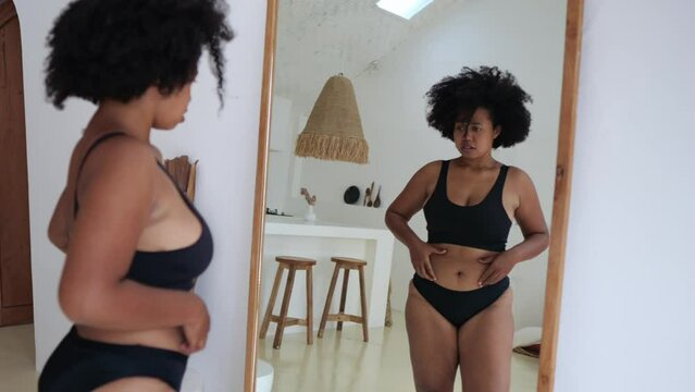 An overweight black woman looks at herself in the mirror and at her fatty parts of the body, the woman has stress due to being overweight. African American woman with obesity.