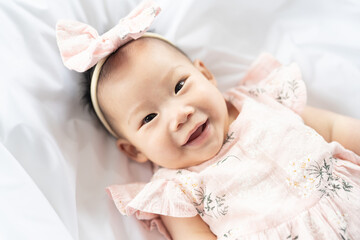 A happy Asian girl playing on the white bed while wearing a pink dress.