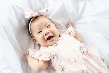 A happy Asian girl playing on the white bed while wearing a pink dress.