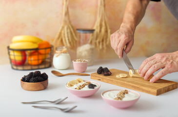 The chef prepares a healthy breakfast or snack. He slices a banana and makes bowls of yogurt, oatmeal and fresh fruit, blackberries. Selectively focused, body parts, hands holding knife.