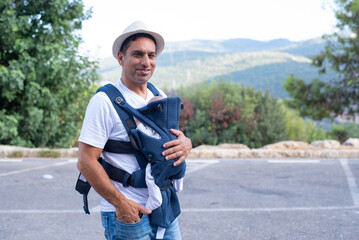 Happy father with hat and with newborn in baby sling standing on background of mountain. Trip to...