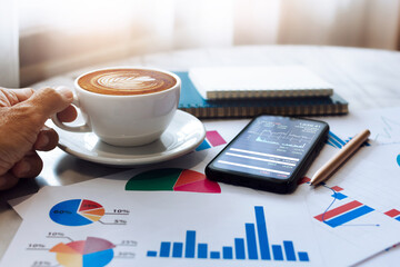 Hand of businessman holding coffee cup with mobile phone, paperwork and chart of investment, business people take a break during a day in cafe after he successful to trading stocks via mobile app