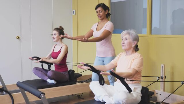 Portrait of a mature woman practicing Pilates in group training with a female instructor, doing exercise on a simulator, sitting in the lotus position. High quality 4k footage