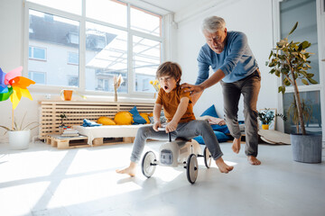 Cheerful grandfather pushing grandson sitting on toy car in living room