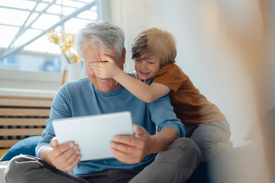 Boy covering eyes of grandfather with tablet PC at home
