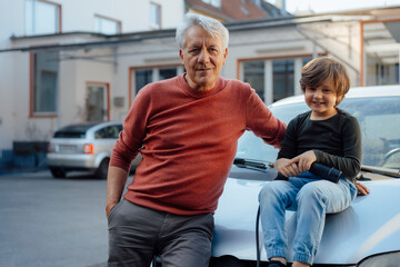Smiling boy with charging plug sitting on car by grandfather