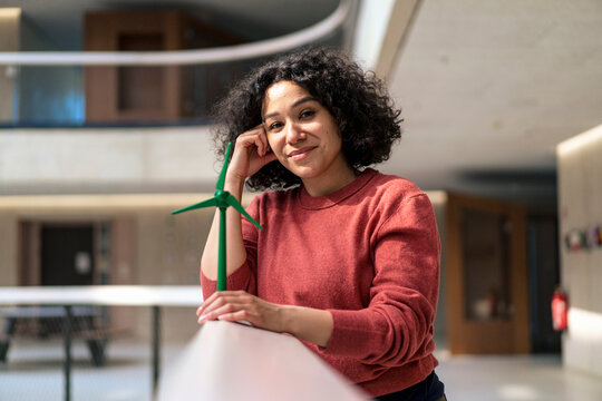 Smiling businesswoman with head in hand holding wind turbine on railing in office corridor