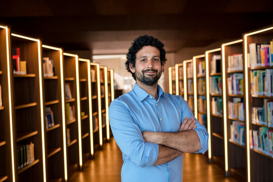 Smiling man with arms crossed amidst bookshelves in illuminated library