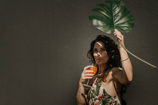 Woman having drink holding monstera leaf in front of wall