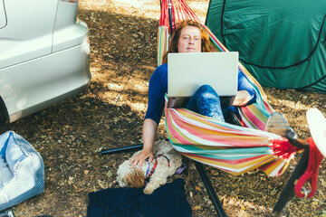 Adult woman working on laptop pc while lying in hammock, stroking cockapoo puppy near motorhome on...