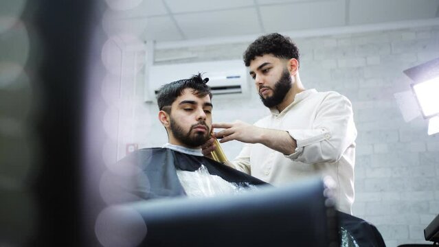 A barber with a beard cuts the man of a client sitting in a chair in a barber shop.