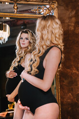 Pregnant beautifull Swedish woman in black bodysuit standing at home looking at large ancient mirror touching her belly, preparing to be a mom. Confident healthy caucasian woman enjoying pregnancy.