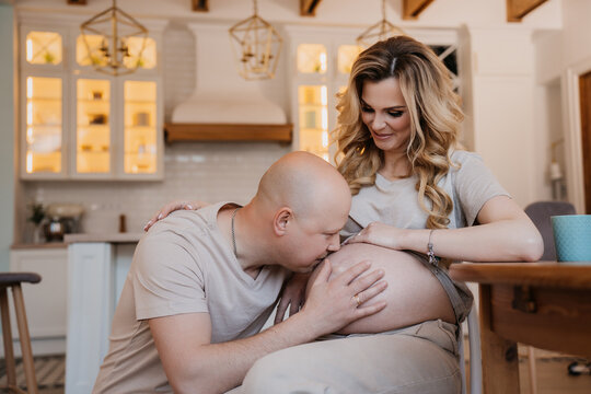 Swedish Mid Adult Bald Man In Beige T-shirt Kissing His Wife’s Belly Putting Head On It Hearing Noises Of A Child Standing On Knees At Kirchen. Happy Caucasian Couple At Home In A Happy Pregnancy.