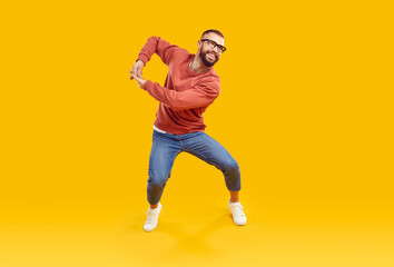 Cheerful male dancer or fashion model in casual clothes dancing and having fun in studio. Happy handsome young man in orange sweatshirt, blue jeans and glasses dancing isolated on yellow background