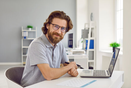 Happy Businessman In Casual T Shirt Working In Office. Handsome Bearded Man In Gray Polo Shirt And Eyeglasses Sitting At Desk With Laptop Computer And Business Calendar, Looking At Camera And Smiling