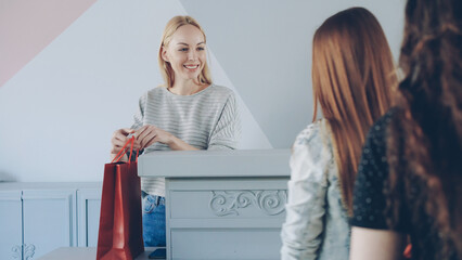 Friendly female cashier is taking garment from customer standing in line folding and packing clothes in paper bag to buyer.