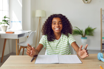 Happy school child sitting at desk at home. Portrait of cheerful African American student girl at table looking at camera and smiling. Online class, videocall or videoconference laptop PC screen view