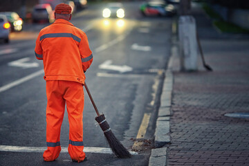 Man sweep road with broom, clean city road and roadside at night. Janitor cleanup city from garbage. Municipal worker in orange uniform sweeping street, night work - obrazy, fototapety, plakaty