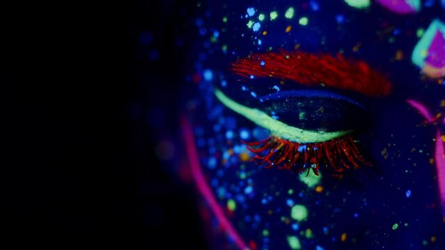 Young woman in bright fluorescent makeup glowing under UV light. Closeup of her eye opening and closing, Studio shot, dark background