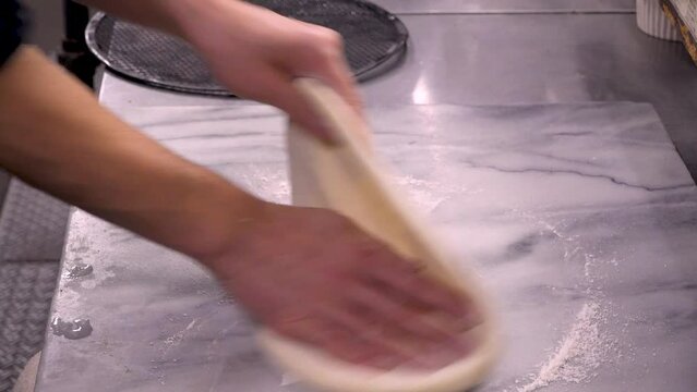 調理台の上でピザの生地をこね、丸く広げていく動作。手際よく生地を押し伸ばし、形を整えていく様子を捉えた映像。飲食店メニュー、イタリアン料理の紹介、自家製ピザや調理工程をテーマにした広告素材に。 | Kneading and shaping pizza dough on a marble counter.