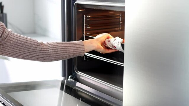 The woman's hands open the oven and pull a baking sheet out of the oven. A young woman in a white modern kitchen cooking food in an oven close up. Concept of Christmas and New Year