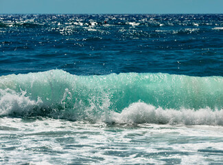 Rough sea with emerald green waves crashing on the beach. In the background the blue sea with sun reflections.