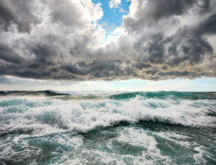 Rough sea with emerald green waves crashing on the beach. In the background the blue sea with sun reflections.