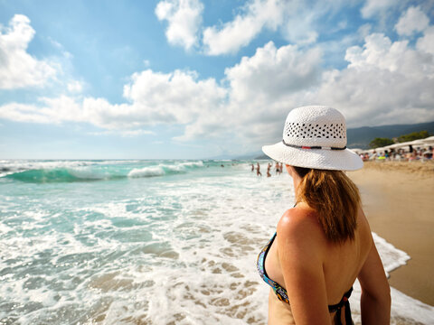 Tanned girl with a white hat, relaxes on the beach looking at the sea on a sunny summer day, in the background bathers cool off by the sea in the Gulf of Policastro, Italy.