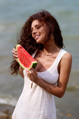 Portrait of a beautiful brunette woman in a white dress at the beach with a slice of watermelon in her hand.
