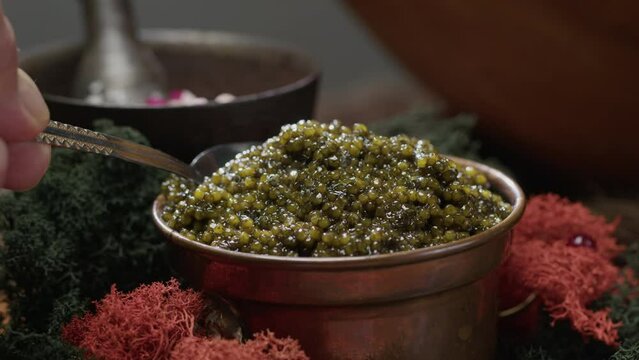 Black caviar in metal dishes is served on dining table. Human hand scoops up teaspoon of delicacy in restaurant, macro close-up