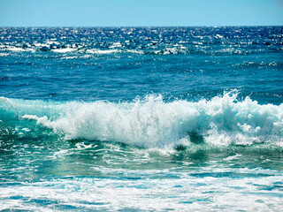 Rough sea with emerald green waves crashing on the beach. In the background the blue sea with sun reflections.