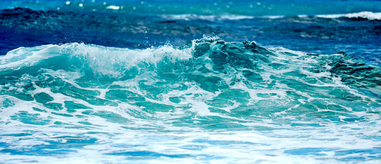 Rough sea with emerald green waves crashing on the beach. In the background the blue sea with sun reflections.