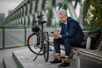 Businessman with bike sitting on bench, using smartphone. Commuting and alternative transport concept