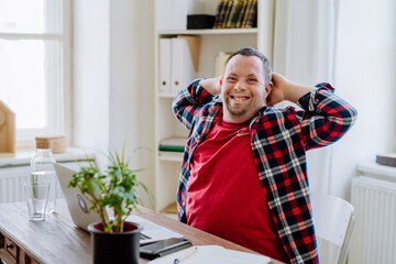 Young man with Down syndrome sitting at desk in office and using laptop, looking at camera and...