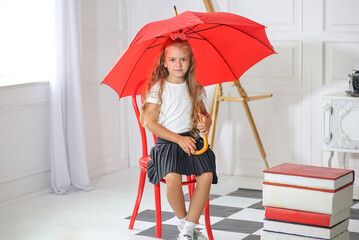 Cute little schoolgirl sitting with a red umbrella on a chair in a room where: table, chair, abacus, books, backpack. Offline and online. Education concept