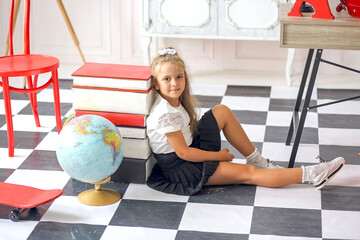 Cute сaucasian girl in a white blouse and black pleated skirt sitting in a school interior. Back to school.Classroom with globe and school supplies.Education concept.