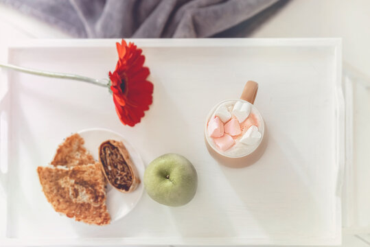 light photo with breakfast, coffee with marshmallows, green apple, red flower and pancakes on a white tray, top view, selective focus