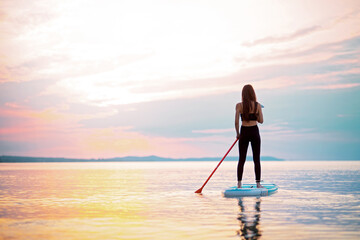 Rear view of girl surfer paddling on surfboard on the lake at sunrise.