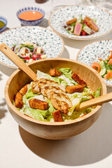 Classic caesar salad with grilled chicken and crouton in wooden bowl on white table. Caesar salad with sauce and wooden spoon. Italian lunch with caesar salad on sunny day. Summer menu.