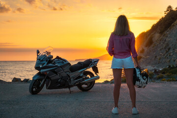 Young woman posing at the beach holds a helmet. Golden sunset and motorbike on the background. Back...
