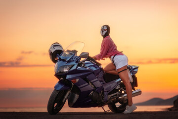 Sexy woman in a black balaclava with skull poses near a tourist motorcycle. Golden sunset in the...